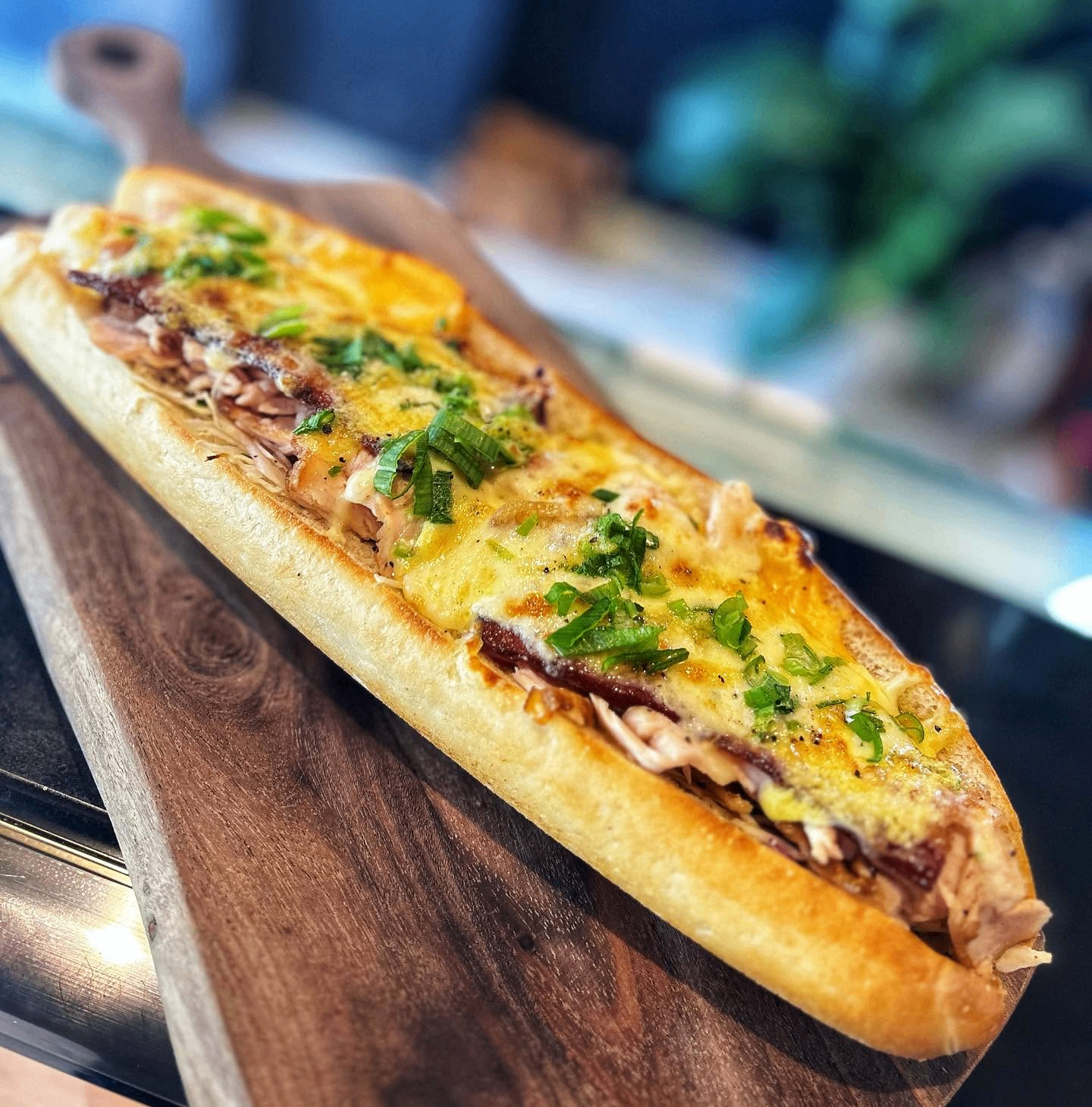 Loaded pulled pork and bacon sub topped with melted cheese and fresh green onions on a toasted roll, served on a wooden board